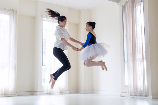 Side view of mother and daughter jumping while performing ballet. Woman and girl are practicing at home. Determined family is dancing against windows.