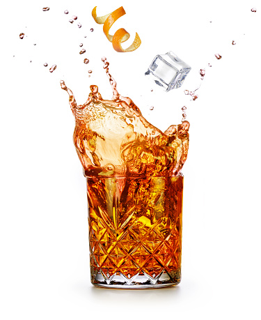Orange zest and ice cube falling into a splashing drink. Godfather or old fashioned cocktail isolated on white background.
