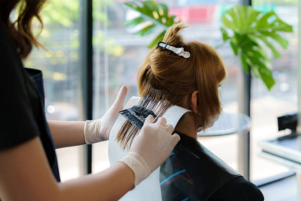 attractive brunette hairdresser thoroughly dyeing hair of female client while she is sitting in chair in beauty salon - matéria corante imagens e fotografias de stock