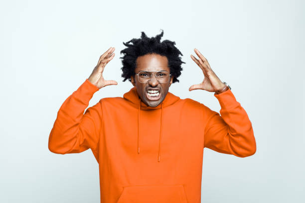 Angry man in orange hoodie Furious afro american young man wearing orange hoodie and glasses, standing with raised hands and screaming at camera. Studio shot on grey background. clenching teeth stock pictures, royalty-free photos & images