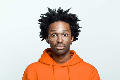Shocked afro american young man wearing orange hoodie and glasses, staring at camera. Studio shot on grey background.