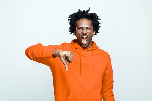 Angry afro american young man wearing orange hoodie and glasses, screaming at camera with thumb down. Studio shot on grey background.