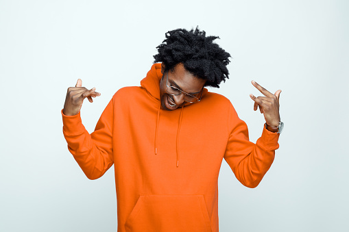 Happy afro american young man wearing orange hoodie and glasses, doing rock symbol with hands up. Studio shot on grey background.