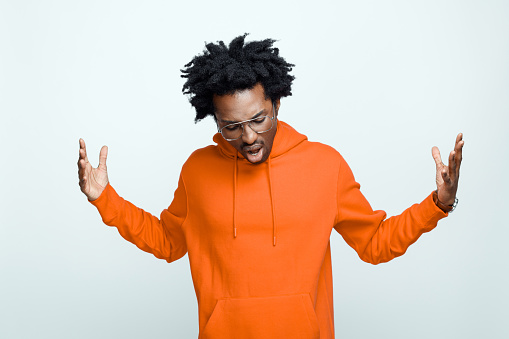 Afro american young man wearing orange hoodie and glasses, raising arms. Studio shot on grey background.