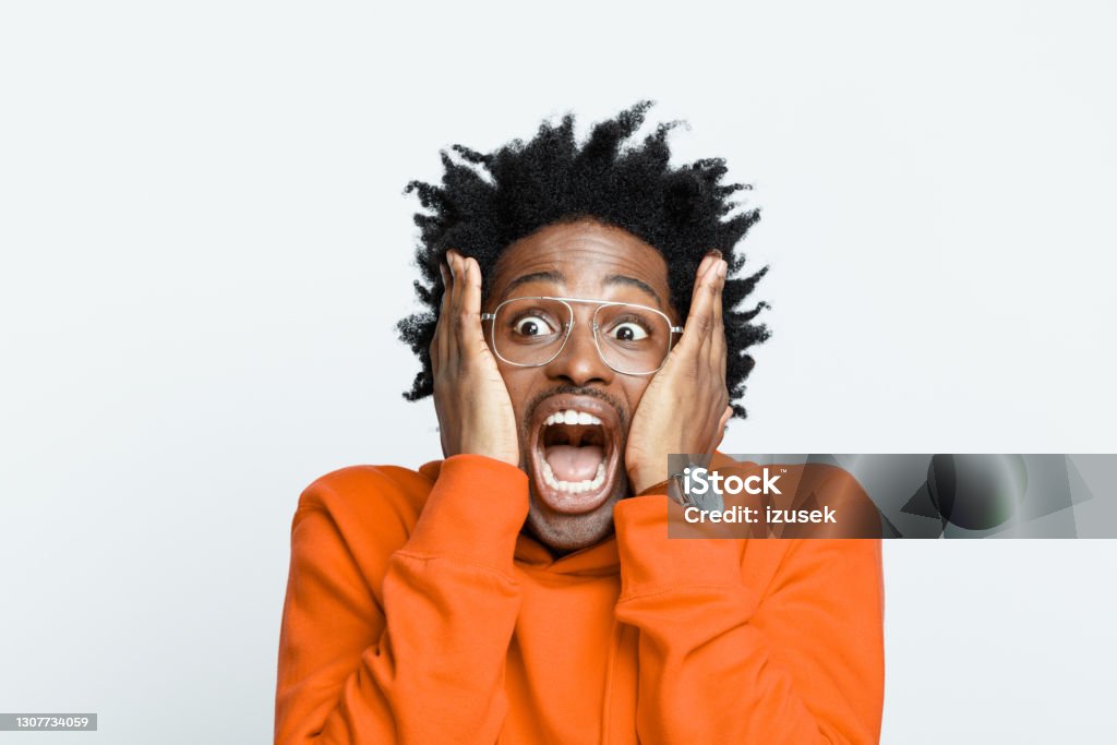 Shocked man in orange hoodie Terrified afro american young man wearing orange hoodie and glasses, looking away and screaming. Studio shot on grey background. Shock Stock Photo