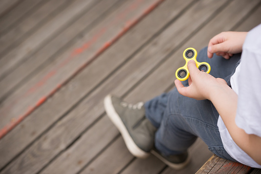 Kid boy sitting on wooden playground and playing with fidget spinner gadget - summer trend of 2017. Yellow hand spinner, fidgeting hand toy rotating on child's hand