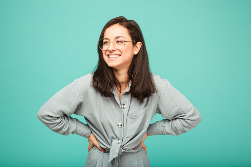 Photo of cute female in glasses smiles nicely. Wears grey shirt, isolated turquoise color background