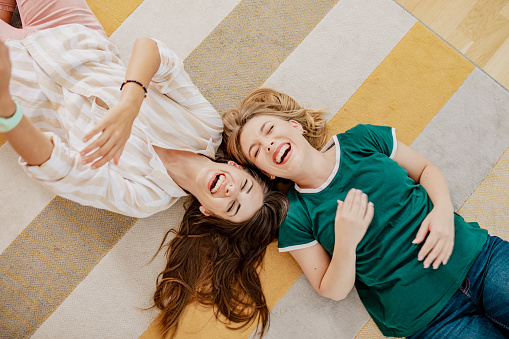 High-angle portrait of two female friends indoors. Two girls are at home on the floor in the living room, they are laughing