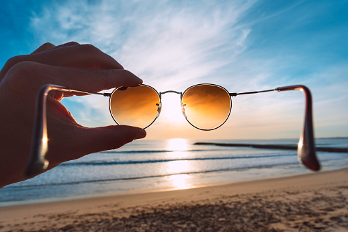 Hand holding stylish round sunglasses with brown lenses at sunset. Putting on sunglasses at sunny summer day near the ocean. Man looking at bright sun through polarized sunglasses. High quality photo
