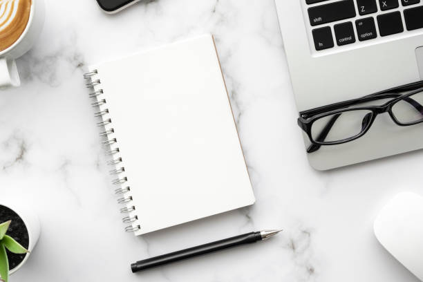 istock Blank notebook with pen are on top of white marble office desk table with laptop computer, cup of coffee and supplies. Top view, flat lay. 1307748286