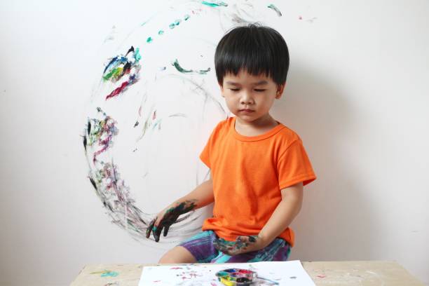 Asian boy select a colour in a paint mixing palette to paint on the concrete wall of the room. little child 3 years old painted on the concrete wall with his hands. stock photo