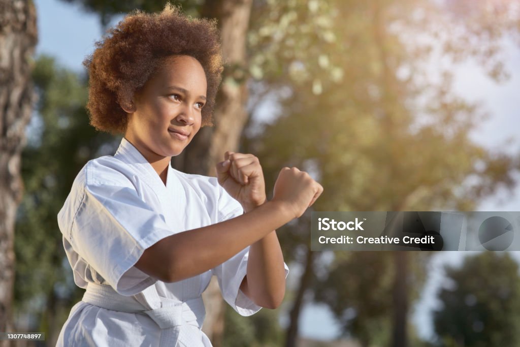 Cute Afro American girl practicing martial arts on sunny day in park Adorable female child karateka looking away and smiling while doing fighting stance on the street Child Stock Photo