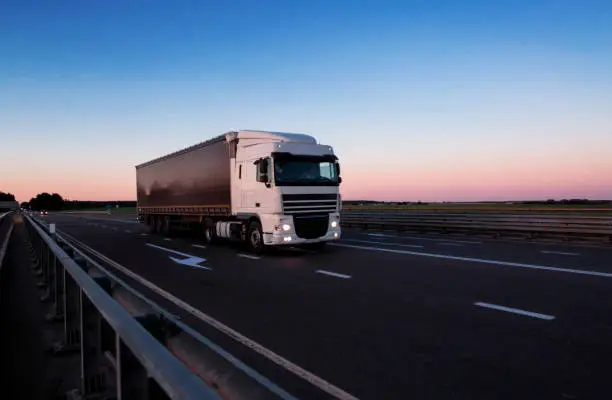 A white semi-trailer truck drives down the highway in the evening with its headlights on. Blue sky and sunset. The mode of work and rest of truck drivers. Copy space for text, transportation
