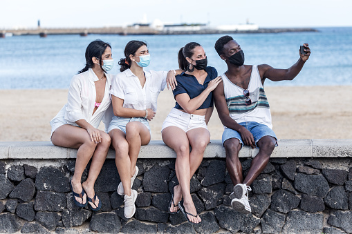Group of diverse friends in masks sitting on stone border near beach and taking selfie on smartphone while enjoying weekend on Lanzarote