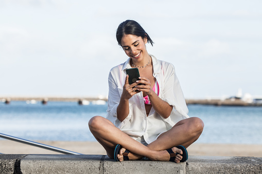 Smiling female in summer clothes sitting with crossed legs on stone border and messaging on smartphone while enjoying weekend on Lanzarote