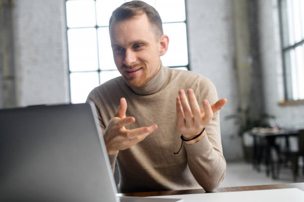 Young handsome businessman in front of a laptop Young handsome businessman entrepreneur sitting at the desk looking at laptop, positive hipster man with the beard pitching the project idea to client or management team online on a video call baseball pitcher stock pictures, royalty-free photos & images