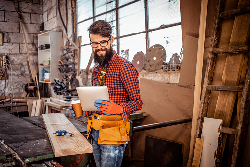 Beautiful young man with beard and glasses in work clothes with protective gloves, cutting wooden board with circular saw in carpentry workshop. He is using tablet to see the project plan and drinking coffee.