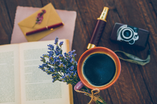 Top view of a coffee mug with lila spring flowers, a book, an old camera, a binoculars, envelope and writing paper in flat lay and boho retro style on dark brown wooden background. Selective focus. Retro editing with added grain. Part of a series.