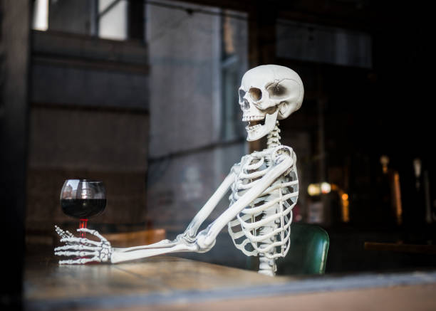 huesos de esqueleto humano sentados en silla de cuero en un pub sosteniendo una copa de vino de alcohol esperando mucho tiempo para que pubs y bares se reabran después de ser cerrados durante el cierre de la pandemia del coronavirus covid-19 - skull holding spooky horror fotografías e imágenes de stock