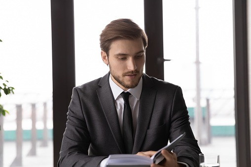 Focused young Caucasian male boss or CEO sit at desk in office look in notebook check plans for day. Serious businessman busy making decisions work with planner or organizer. Time management concept.