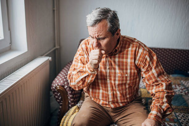Ill senior man coughing One man, worried senior man, sitting alone at his home. He has a pain in his knee. coughing stock pictures, royalty-free photos & images