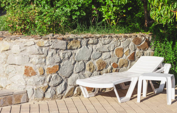 istock White plastic chaise lounge and table on a tiled surface next to a stone wall under green trees on a bright sunny day 1307803339