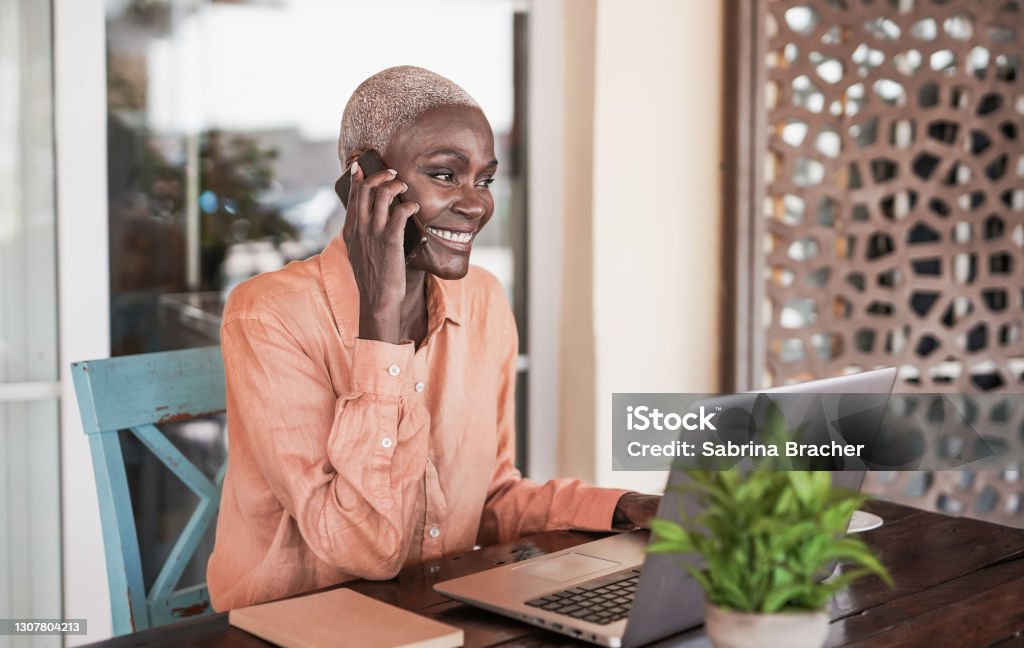 Mature black woman working with laptop outdoor while talking on smartphone call One Woman Only Stock Photo