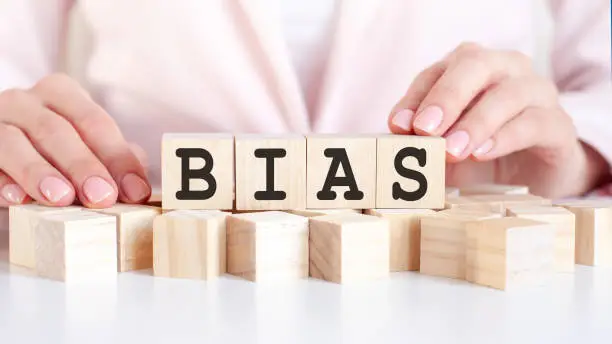 The hand puts a wooden cube with the letter Bias. The word is written on wooden cubes standing on the white surface of the table on the pink background.