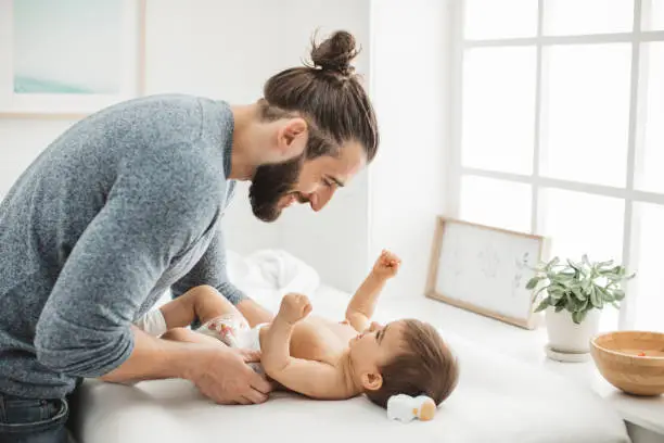 Young father playing with his baby girl while changing diaper on baby changing table