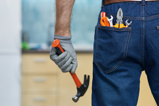 Nail it. Close up shot of various plumbing hand tools in man's jeans back pocket. Professional repairman holding a hammer, standing indoors. Repair service concept