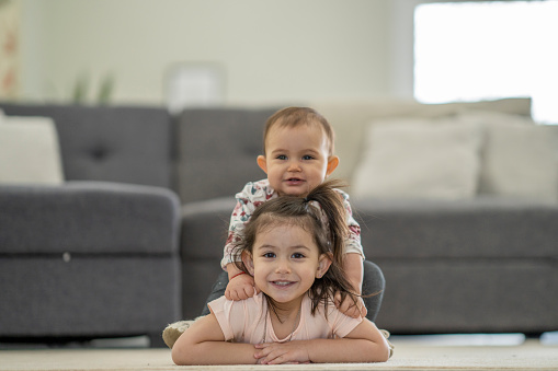 A 3 year old girl is laying on the floor and her baby sister is sitting her back and hugging her from behind. They are both happy and smiling at the camera.