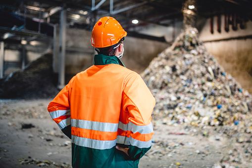 Rear view of young male worker in helmet, pollution mask, and reflective clothing observing waste falling from conveyor belt onto pile at facility.