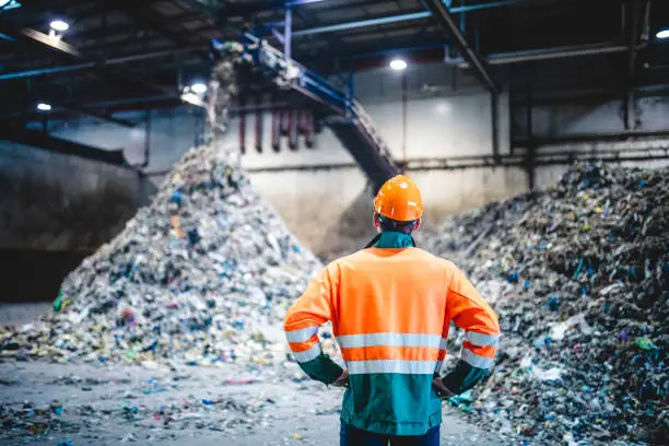 Rear view of young male worker in helmet, pollution mask, and reflective clothing observing waste falling from conveyor belt onto pile at facility.