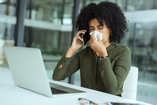 Shot of a young businesswoman blowing her nose while using a laptop and smartphone in a modern office