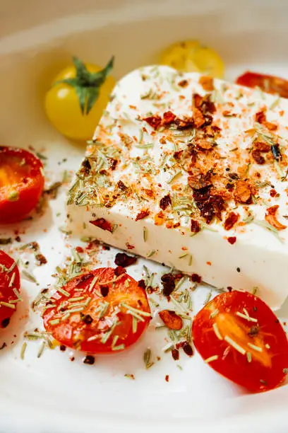 Photo of baked feta cheese with small tomatoes in olive oil with herbs and spices in ceramic baking dish on table with yellow cotton kitchen towel. popular vegan recipe home cooking. eat less meat. top view
