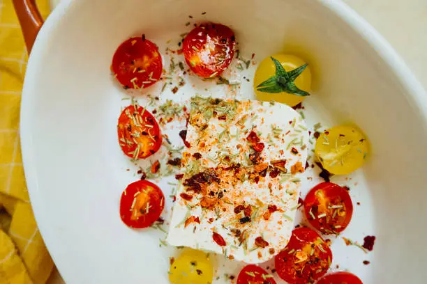 Photo of baked feta cheese with small tomatoes in olive oil with herbs and spices in ceramic baking dish on table with yellow cotton kitchen towel. popular vegan recipe home cooking. eat less meat. top view