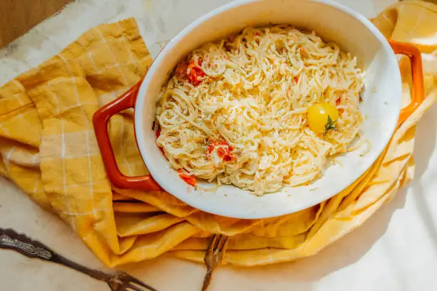 Photo of pasta from feta cheese with small tomatoes in olive oil with herbs and spices in ceramic baking dish with yellow cotton napkin. popular vegan recipe home cooking. eat less meat. top view