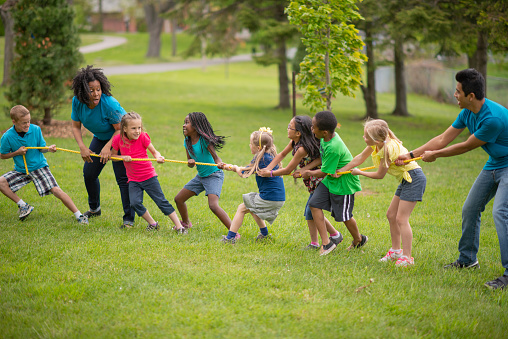 A group of children are playing tug of war outdoors at a summer camp. Their camp counsellors at helping them.