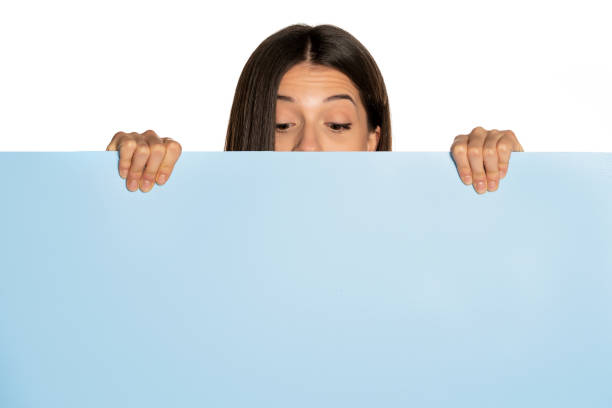 istock young beautiful woman peeking behind a blue empty board on white background 1307843609