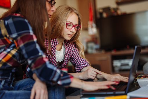 Mother helping daughter in searching information for homework on internet or attending online class while sitting together on sofa with laptop at home