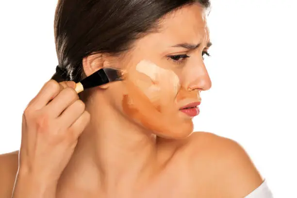 Photo of Young nervous woman smearing three different tones of liquid foundation on her face on white background