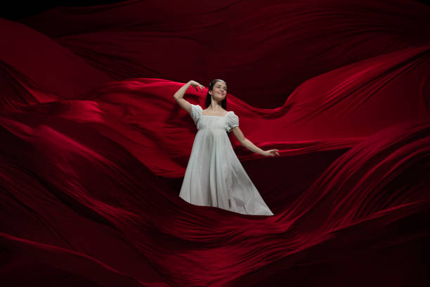 istock Young and graceful ballet dancer on billowing red cloth background in classic action. Art, motion, action, flexibility, inspiration concept. 1307852263