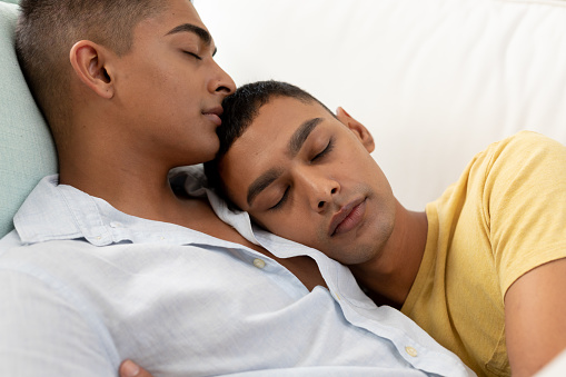 Diverse gay male couple sleeping on sofa and embracing. staying at home in isolation during quarantine lockdown.