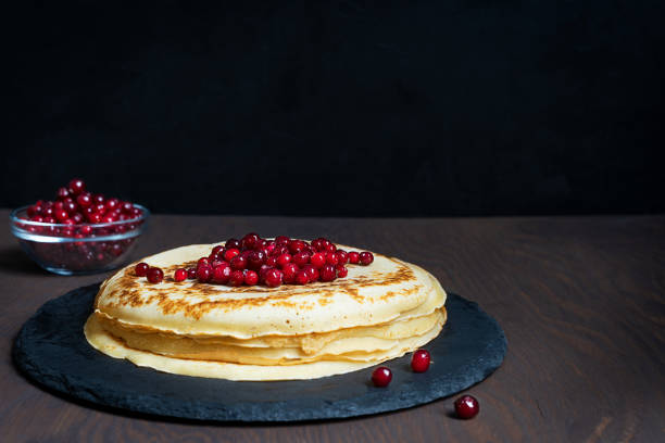 Pile of russian homemade traditional blinis, also crepes with red berries served on round stone tray ready for eating on dark brown wooden table against black background. Image with copy space