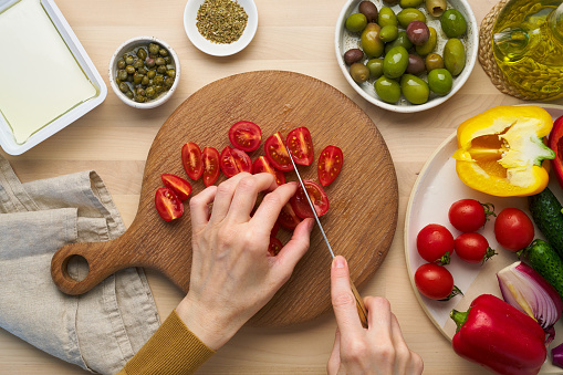 Vegetarian vegan food. Chopping tomatoes, cutting vegetables for greek salad horiatiki. Woman hands with knife on wooden cutting board slicing chop of vegetables. Homemade vegan food, top view