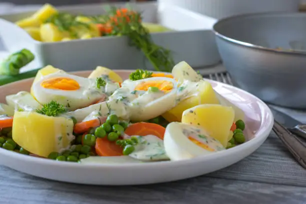 Delicious and cheap meal with boiled, halved eggs, cooked potatoes and peas and carrot vegetables served with a herb bechamel sauce on a plate.