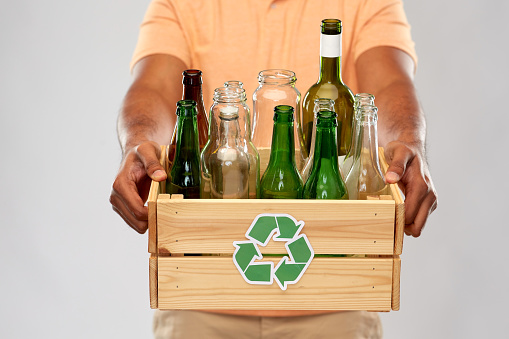 recycling, waste sorting and sustainability concept - close up of african american young man holding wooden box with glass bottles and jars over grey background