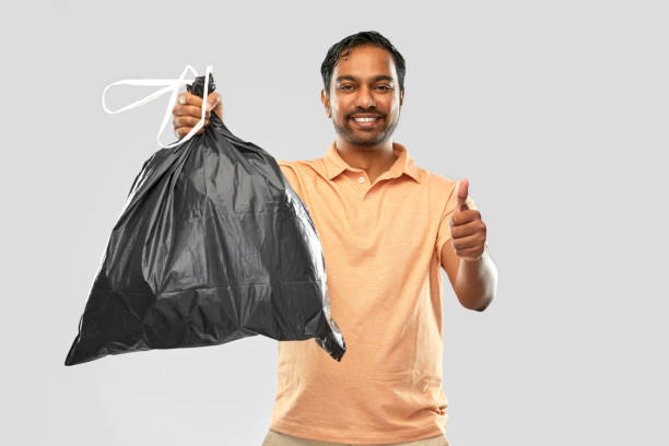 recycling, sorting and sustainability concept - smiling young indian man holding trash bag and showing thumbs up over grey background