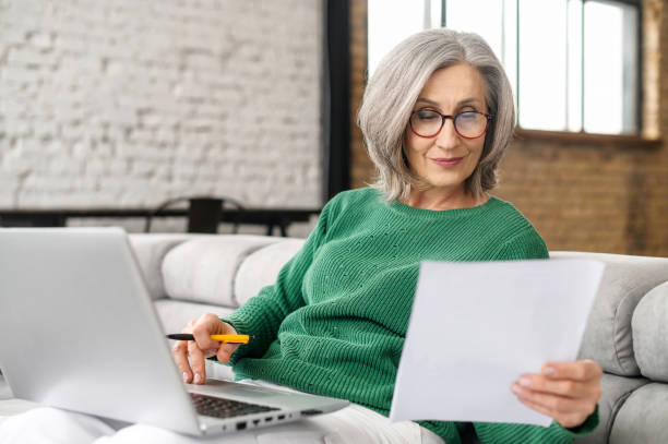 Mature senior woman using a laptop at home Confident elderly businesswoman is doing paperwork sitting with a laptop on the couch at home, focused senior employee preparing documents, checking bills working remotely at home beak stock pictures, royalty-free photos & images