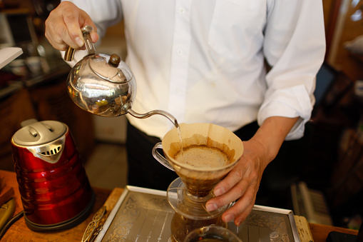 asian male barista pouring boiling water from kettle to drip coffee maker on wooden table